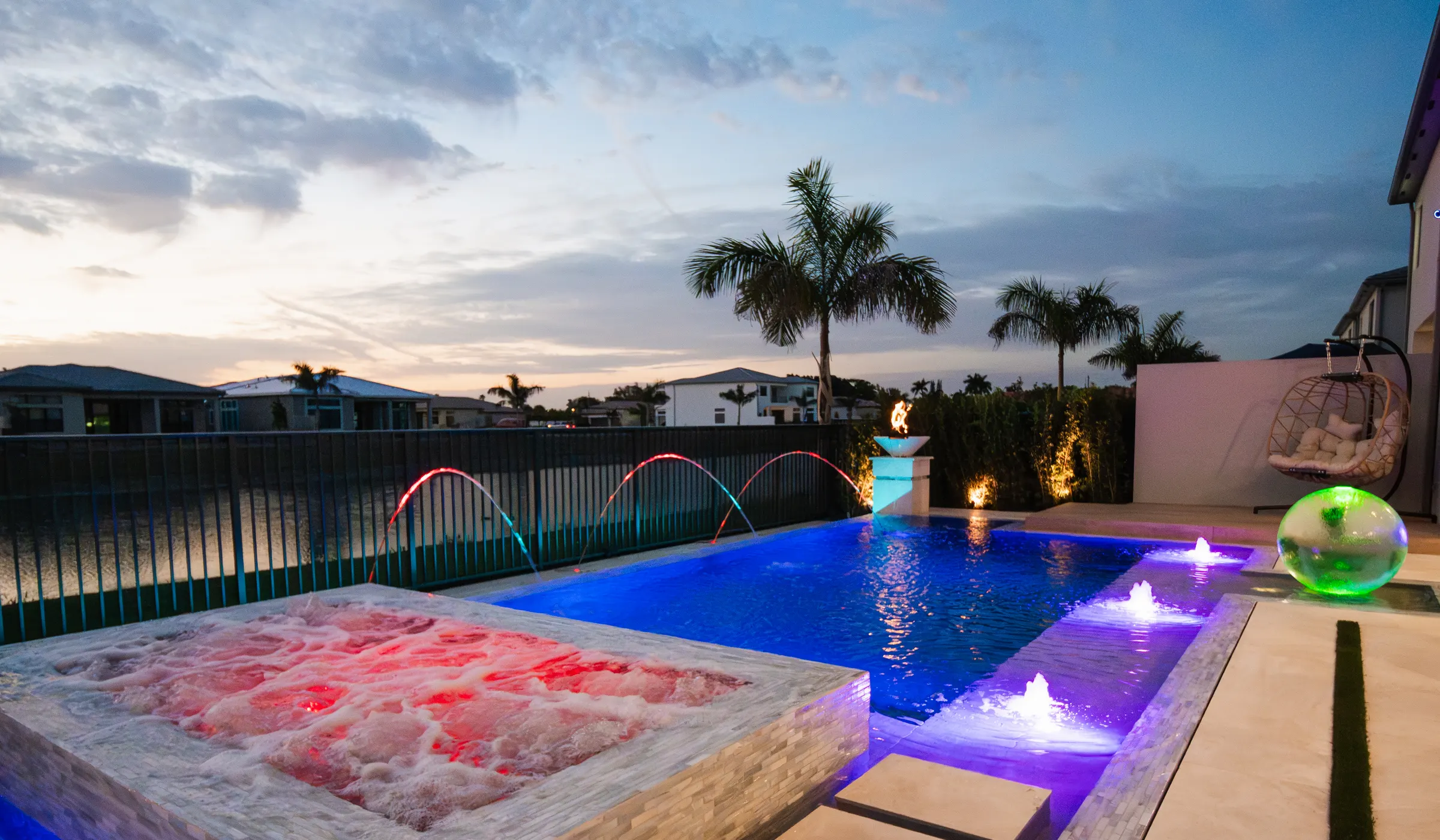 Backyard pool and spa at dusk under a dramatic sunset sky, with a linear fire feature, colored water jets, and palm trees at a Liquid Luxe project in South Florida